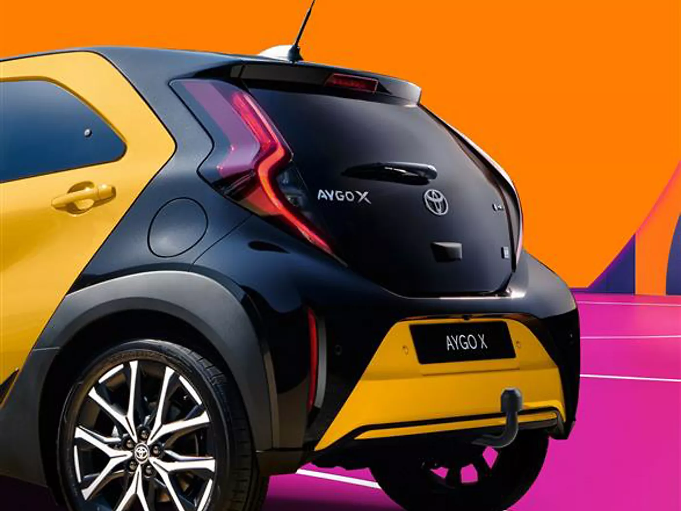 Nahaufnahme des Hecks eines schwarz-gelben Toyota Aygo X vor einem orangen Hintergrund. Das Fahrzeug steht auf einem pinken Untergrund mit weißen Linien.
