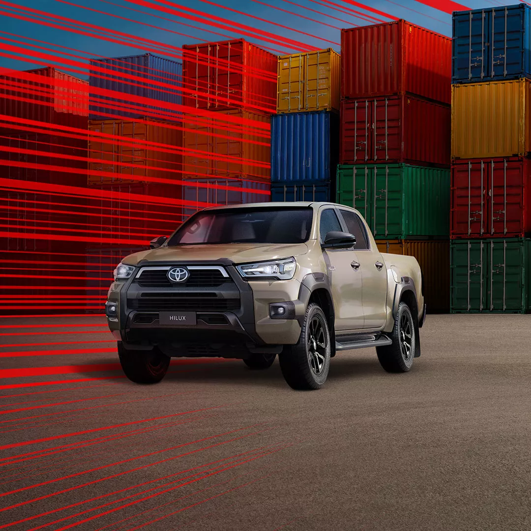 Ein grau-brauner Toyota Hilux steht vor einer Lagerhalle, daneben stehen Holz-Paletten.