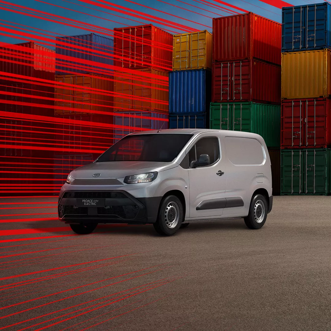 Ein grauer Proace City Electric steht vor einem gläsernen Gewächshaus neben einer Ladesäule und wird geladen.